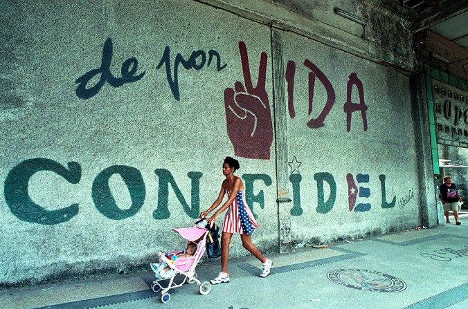 Une femme vêtue aux couleurs du drapeau américain passe avec son bébé devant un mur où on lit : « Toute la vie avec Fidel », le 11 juin 2001, à  La Havane, Cuba. (Jorge Rey / Getty Images)