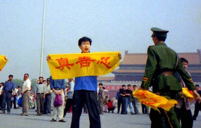 Un policier chinois s'approche d'un pratiquant de Falun Gong sur la place Tiananmen à Pékin alors qu'il tient une bannière avec les caractères chinois signifiant « Authenticité, Compassion et Tolérance », les principes fondamentaux du Falun Gong (via Minghui.org)