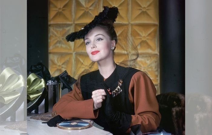 En 1945, l'actrice et mannequin américaine Anita Colby, assise à une coiffeuse entourée de boîtes à chapeaux, sourit et enlève ses gants. Elle porte un ensemble noir et roussâtre ainsi qu'un chapeau noir et un voile en filet. (Hulton Archive/Getty Images)