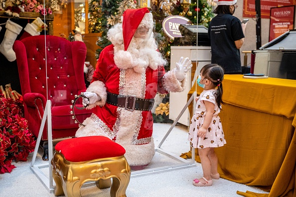 -Un homme vêtu d'une tenue de père Noël, portant un écran facial est agenouillé derrière une barrière transparente, fait signe à une fille dans un centre commercial à Kuala Lumpur le 22 décembre 2020. Photo de Mohd RASFAN / AFP via Getty Images.