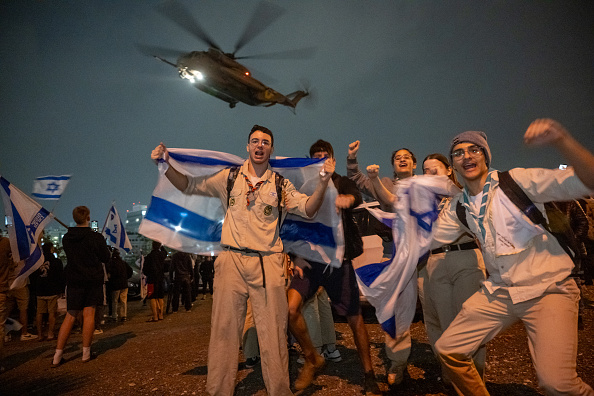 Des groupes de jeunes israéliens agitent des drapeaux. Au-dessus d'eux, un deuxième hélicoptère transportant des otages libérés, le 26 novembre 2023 à Petah Tikva, Israël. (Photo Alexi J. Rosenfeld/Getty Images)