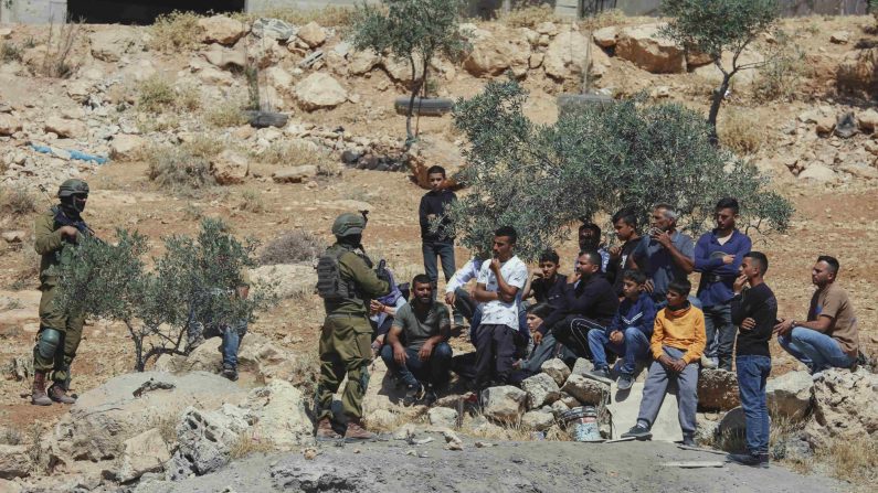 Des soldats israéliens se tiennent près d'un groupe de résidents palestiniens assis dans le quartier Al-Rifaiya de Yatta, au sud d'Hébron, en Cisjordanie, le 28 mai 2025. Les forces d'occupation démolissent la maison du Palestinien Jihad Al-Amour, qui abritait une famille de sept personnes, dont une mère et quatre enfants, sous prétexte de construction sans permis dans la zone C. (MOSAB SHAWER/Middle East Images/AFP via Getty Images) 